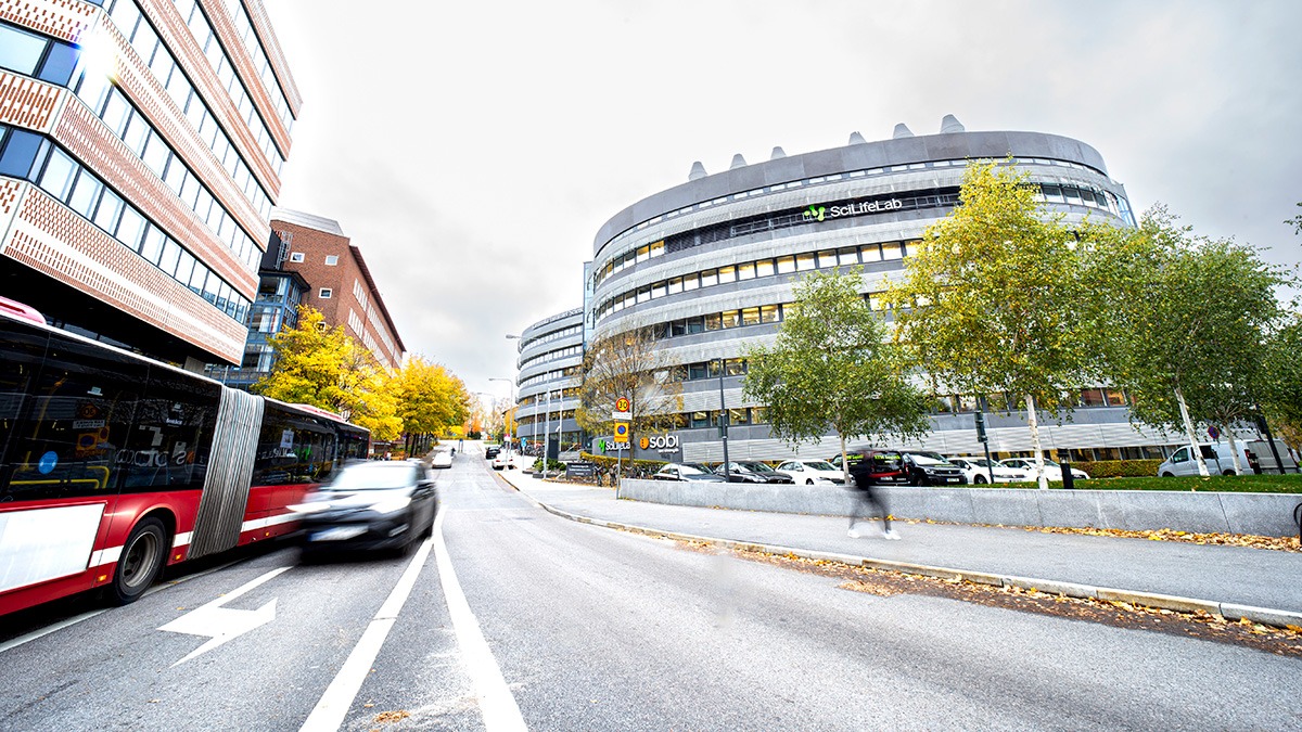 The Campus Solna building with the SciLifeLab logo visible. Photograph taken from the road next to the buildings. A bus, car and person on bike are speeding by and are blurry.