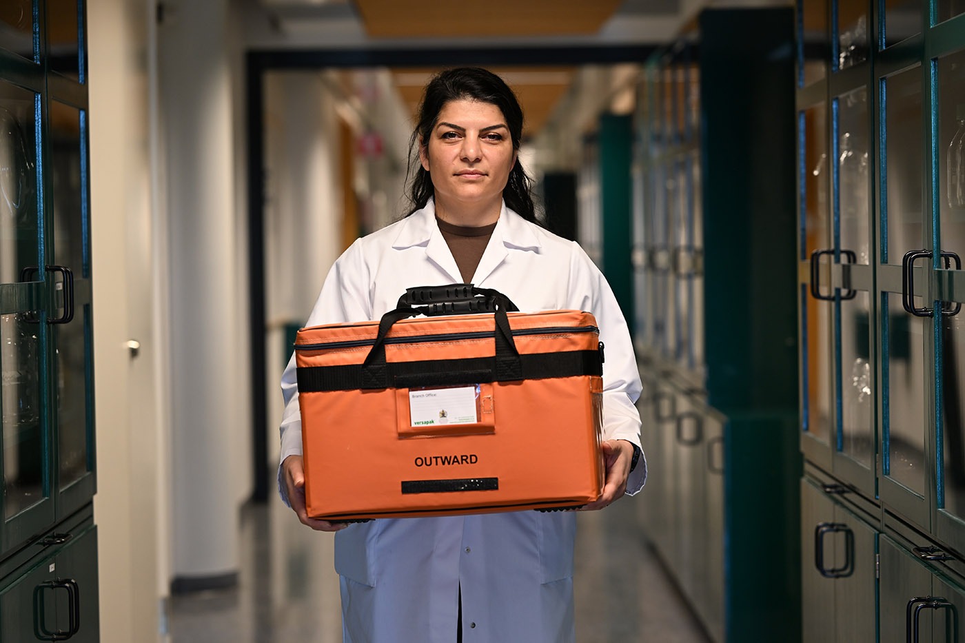 Ghazaleh Assadi stands in in the middle of a corridor with glass covered shelves. Ghazaleh looks into the camera, holding an orange bag to transport samples.